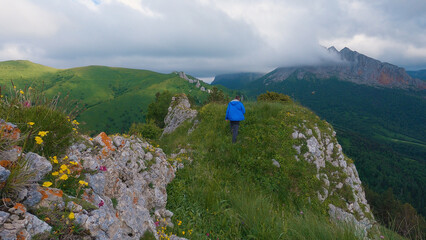 A lone figure in a blue jacket strides across a rocky terrain surrounded by the greenery of the mountainous landscape. An overcast sky looms overhead, white clouds kissing peaks of distant mountains.