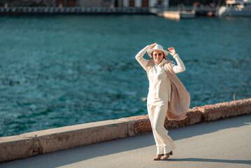 Happy blonde woman in a white suit and hat posing at the camera against the backdrop of the sea