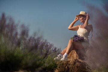 woman sitting in a field of lavender and wearing a straw hat. She is smiling and holding a bouquet of flowers. Scene is peaceful and serene, as the woman is surrounded by the beauty of nature