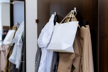 Mockup of white paper bag hanging with clothes on a hanger in the locker room in clothing store, in...