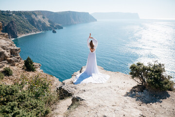 Fototapeta na wymiar A woman stands on a cliff overlooking the ocean in a white dress. She is looking out at the water and she is in a state of peace and serenity. Concept of calm and tranquility.