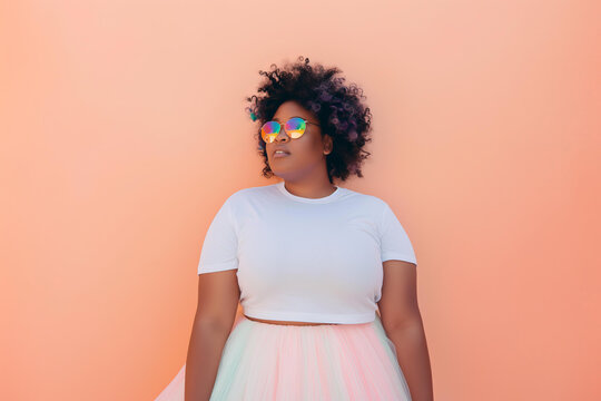 plus sized young woman wearing holographic sunglasses blank white tshirt and tulle skirt against a pastel peach backdrop