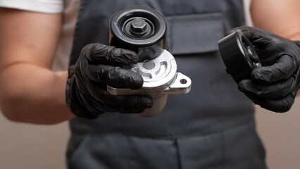 An auto mechanic holds in his hands in black gloves a idler pulley with a tensioner for a car's poly V-belt and a parasitic roller, close-up. Auto parts replacement concept, car maintenance
