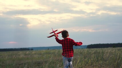 Casual playful boy kid running with red plane pilot flying at evening field dramatic sky back view slowmo. Adorable male child playing airplane flight fantasy imagination enjoy freedom happy childhood