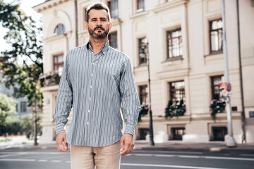 Portrait of handsome confident stylish hipster lambersexual model. Sexy modern man dressed in blue shirt and trousers. Fashion male posing on street background in Europe city at sunset, sunglasses