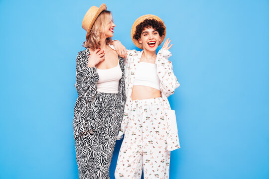 Two young beautiful smiling brunette hipster female in trendy summer costume clothes. Sexy women posing near blue wall in studio. Positive models having fun. Cheerful and happy. In hat