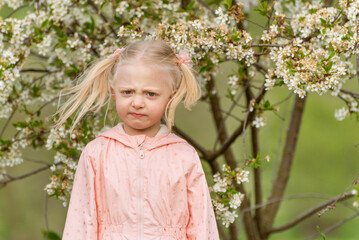 Portrait of frustrated or angry little girl outside near flowering tree with funny grimace on her face.