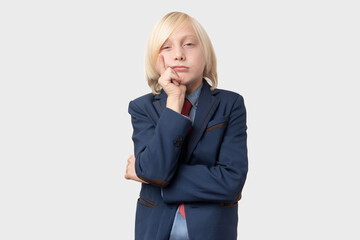 Portrait of pensive blonde boy with short hair, keeps hand on chin, wears formal suit, isolated over white background. People sincere emotions, lifestyle concept.