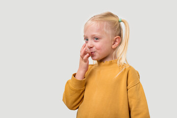 Cute little girl with blond hair in yellow sweater making funny face and looking at camera while standing against grey background