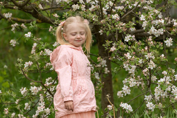 Cute blond preschool girl with closed eyes wears light pink jacket stand near flowering tree in spring garden. Spring time