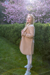 Thoughtful middle-aged woman in beige coat with long blonde hair in spring park. Full-length portrait . Vertical frame.