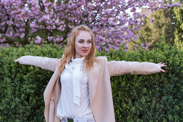 Portrait of scarlet-haired woman with curly hair with raised arms in blooming spring park. Girl on blooming sakura tree background.