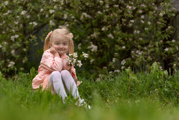 Funny little girl in pink dress and white tights sits on grass with fist under her chin and looks at the camera in surprise.