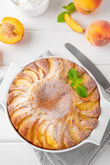 Delicious peach pie in a white ceramic baking form on a white wooden background. Summer cake. Selective focus, copy space.