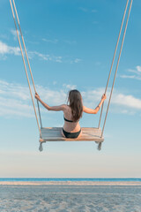 Young slim girl in swimsuit pushes on rope swing on blue sky and beach background. Vertical frame.