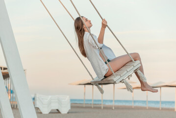 Happy beautiful young girl in white clothes pushes in swing on sky background. Beautiful soft sunset light. Pretty girl on the beach