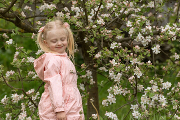 Cute flirty preschool girl with closed eyes wears light pink outfit standing near flowering tree in spring garden. Spring time