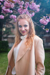 Portrait of attractive young woman in stylish beige coat near blossoming pink tree in park in early spring. Fashion model with red wavy hair.