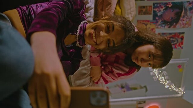 Vertical video of two multicultural teenage girls in the cozy bedroom taking selfie pictures using mobile phone. Happy best friends spend leisure time at home. Beautiful besties having fun together.
