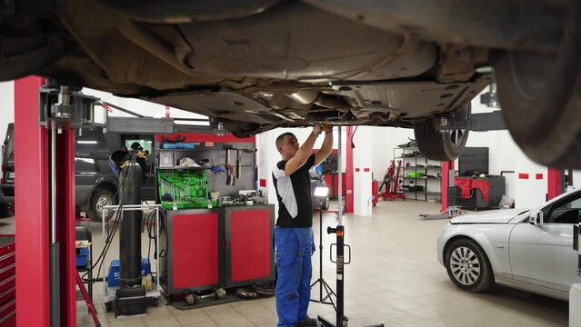 Auto mechanic installs gearbox in vehicle at repair shop. Worker fits transmission, conducts clutch maintenance under lifted car. DIY car enthusiast updates auto parts, restores in garage.