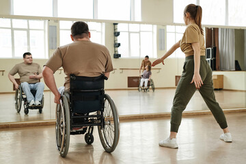 Rear view of man in wheelchair looking at young female dance trainer while she demonstrating moves to him