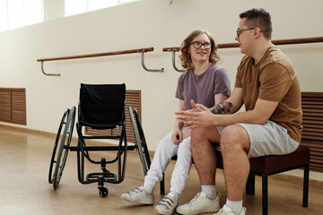 Fototapeta premium Long shot of modern Caucasian female wheelchair dancer and her partner sitting on bench in dance studio chatting during break