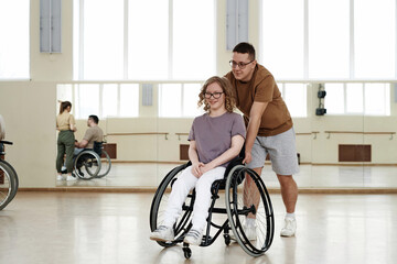 Long shot of of young man pushing wheelchair with cheerful young woman in it during workout in...