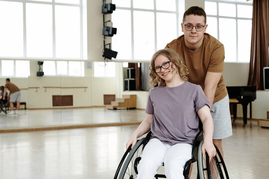 Medium long portarit of cheerful young woman in wheelchair and young man standing behind her in dance studio, copy space - Powered by Adobe