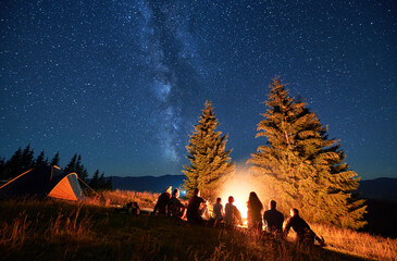 Night camping in mountains under starry sky. Group of people hikers having a rest near campsite, burning campfire and tourist tent. Concept of tourism, hiking and adventure.