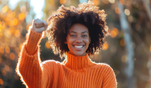 Black woman celebrating with fist in the air, smiling and wearing an orange sweater.