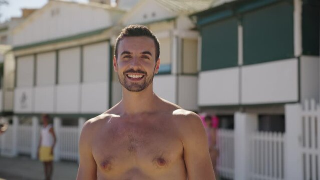Young Caucasian handsome man in swimwear smiling at camera on sunny summer day at beach. Happy European attractive male of generation z posing joyful with positive attitude on vacation outdoors