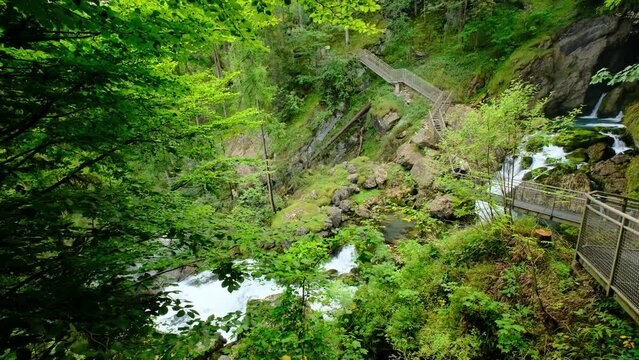 A beautiful, green lush river, where the stream comes out of a cave in the mountain, and runs below a twisted trail deck. few unrecognizable people are walking on the path in the distance. 4K video.