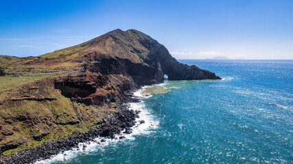 
The rocky coast of Madeira, with green cliffs rising above the blue ocean. Waves gently crash against the base of the cliffs, creating white foam. The sky is almost clear with a few clouds.