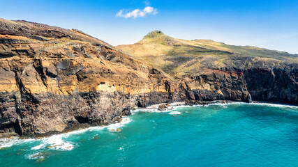 The rocky coastline with a light blue sea, rocks, and green hills. It's a sunny day. PR8 trail in...