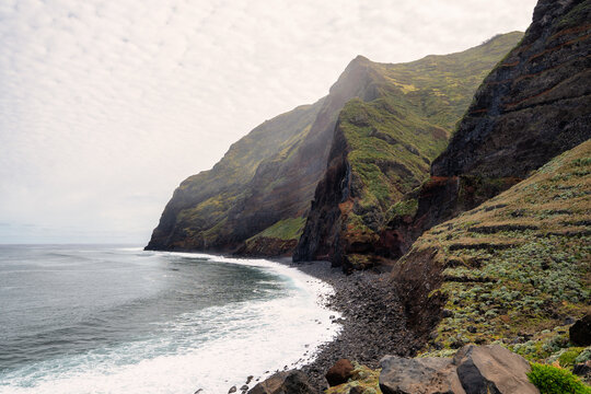 The rocky coast of Madeira, as seen from the Achadas da Cruz viewpoint. Green slopes, a white beach, and blue sea are visible under a partly cloudy sky. The overall mood of the picture is calm and