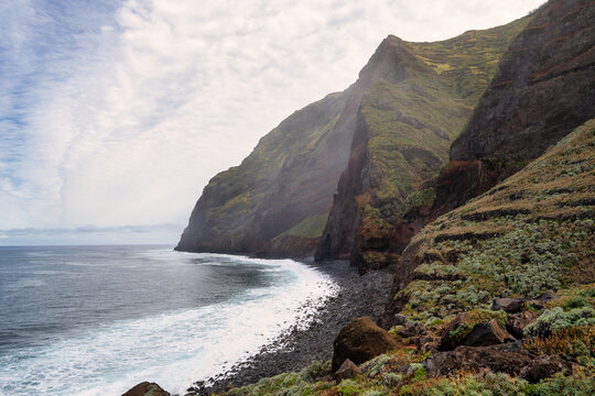 The photo depicts a steep, green mountainside in Madeira, sloping down to the blue sea. Waves crash against the rocky coast, and dense clouds dominate the scene, creating an image of untouched nature.