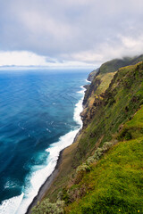 The steep, green cliffs of Madeira above the azure ocean. Mist envelops the peaks, and waves crash against the rocky coastline. It's the beauty of nature and the rugged charm of the island.
