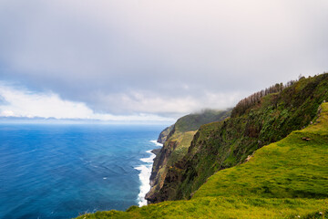 The steep, green cliffs of Madeira above the azure ocean. Mist envelops the peaks, and waves crash...