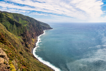 The photo depicts a steep, green mountainside in Madeira, sloping down to the blue sea. Waves crash...