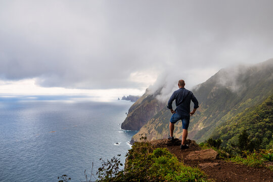 The picture shows a person standing at the edge of a cliff, overlooking the ocean and the mountains of Madeira. The mountains are steep, covered in greenery, and partially shrouded in mist.