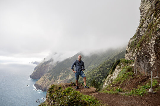 The picture shows a person standing at the edge of a cliff, overlooking the ocean and the mountains of Madeira. The mountains are steep, covered in greenery, and partially shrouded in mist.