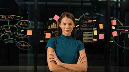Fototapeta na wymiar Professional businesswoman looking at camera while standing with arms crossing. Skilled beautiful worker crossing arm while standing in front of glass wall with mind map and sticky notes. Tracery