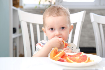 A little cute child boy is eating orange in light kitchen. 