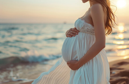A pregnant woman stands on the beach, wearing white and holding her belly with both hands.