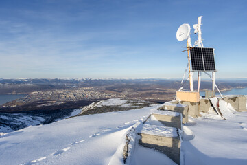 Solar panels, antennas and CCTV camera (webcam) on a snowy mountain top. In the distance is a large northern city. Neighborhoods of Magadan, Magadan region, Russian Far East.