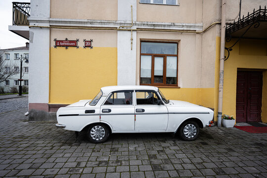 Lutsk, Ukraine - March, 2024: A classic white soviet Moskvitch sedan car carefully parked by a colorful building facade with traditional European architectural elements and cobblestone pavement.