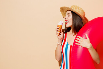 Side view young sexy woman wear one-piece swimsuit straw hat near hotel pool hold pink inflatable rubber ring eat icecream isolated on plain beige background. Summer vacation sea rest sun tan concept.