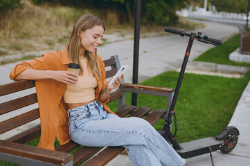 Side view happy fun young woman wear shirt casual clothes use mobile cell phone drink coffee sit on bench near e-scooter walk rest relax in spring city park outdoors on nature Urban lifestyle concept. - obrazy, fototapety, plakaty