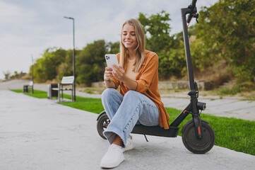 Full body young happy woman wears orange shirt casual clothes sit on e-scooter using mobile cell phone chatting walking resting relax in spring city park outdoors on nature. Urban lifestyle concept.