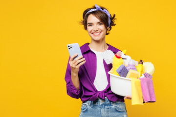 Young happy woman wear purple shirt hold basin with detergent bottles do housework tidy up hold in...